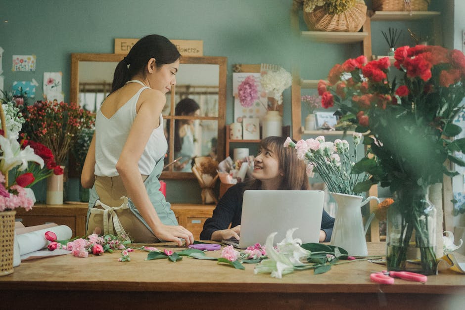 A florist and a customer inside a florist shop engaged in a conversation about floral arrangements. The florist, a young woman with dark hair tied back, is standing at a wooden worktable, wearing a wh