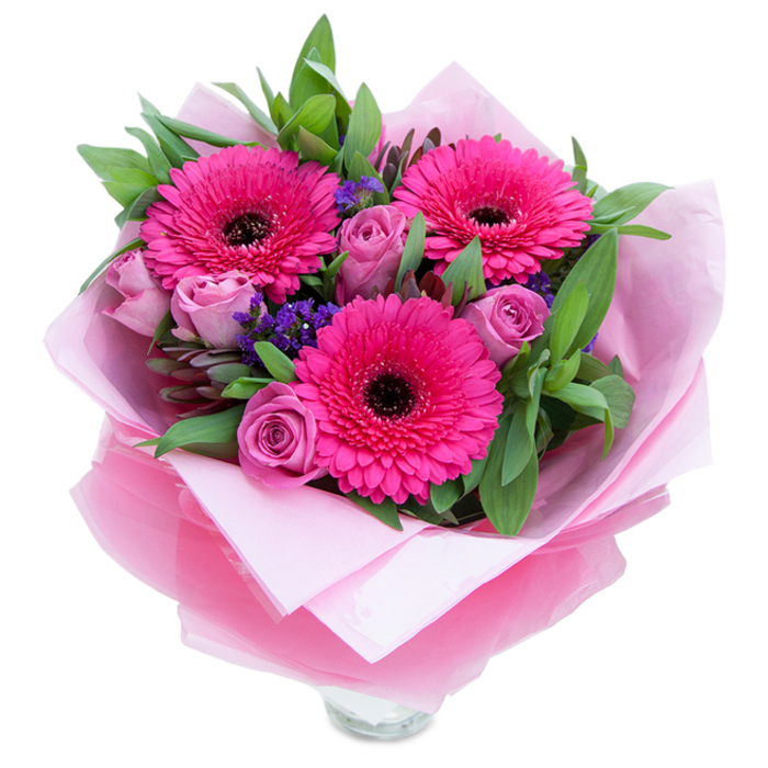 A tender, expertly composed bouquet that blends playful colour with graceful form, centred on three vivid fuchsia Gerbera daisies whose layered petals open like small suns around deep centres. Nestled beside them are soft lavender-pink roses, their petals still holding the gentle spiral of young buds and offering a subtle, classic rose scent that mingles with a faint green sweetness from the gerberas. Little sprays of purple statice weave through the arrangement, their fine texture catching light and giving the composition an informal, meadow-like rhythm. The greenery-broad salal leaves and a few eucalyptus stems-wraps the flowers in a fresh, slightly resinous fragrance and gives visual balance with glossy, cool-toned foliage. The bouquet is presented wrapped: an inner layer of pale pink matte tissue creates a soft, protective cradle, while an outer sheet of translucent fuchsia-pink cellophane adds a sheen and tactile crinkle, making the gift feel celebratory. Visible at the base is the upper rim of a clear glass vase, indicating the flowers are arranged for immediate display on a hallway table or kitchen island. In the gentle morning light, the pinks feel both bright and intimate-ideal for marking birthdays, Mother's Day, anniversaries, or a thoughtful surprise for someone across Cranford Park or the high street. The overall effect is one of considered craftsmanship: bold focal blooms tempered by delicate roses and airy statice, arranged to look effortless and to lift the spirits of the recipient.